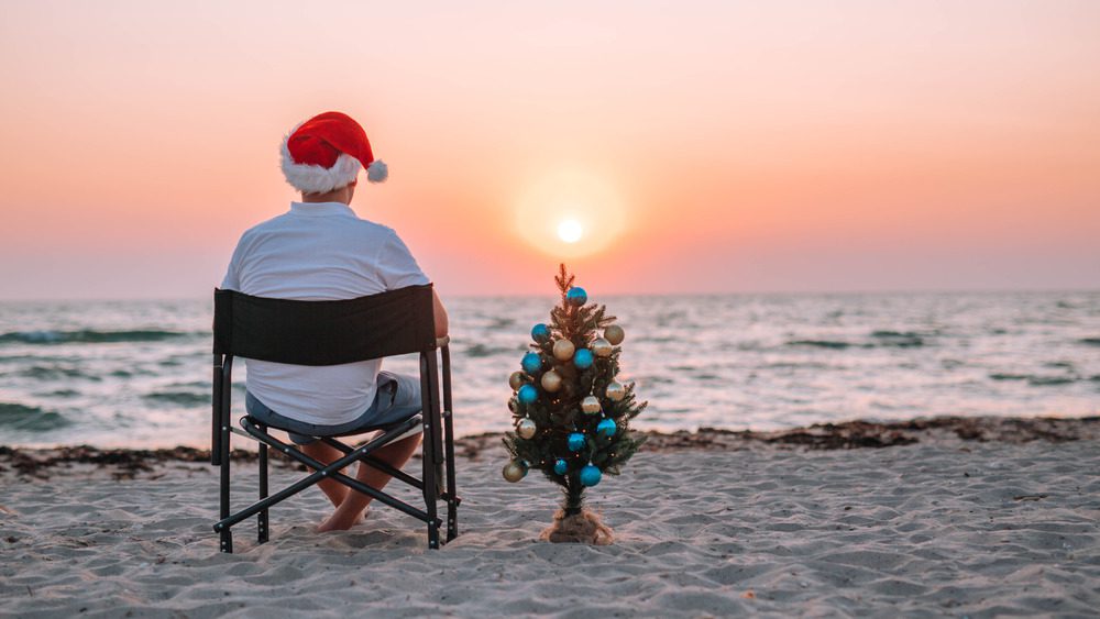 Мужчина сидит с рождественской елкой на пляже.